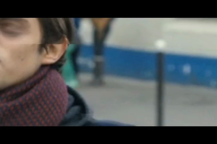 Photogramme : le profil flou d'un homme en écharpe grenat et manteau sombre, dans une rue où passent des barrières bleues et blanches.