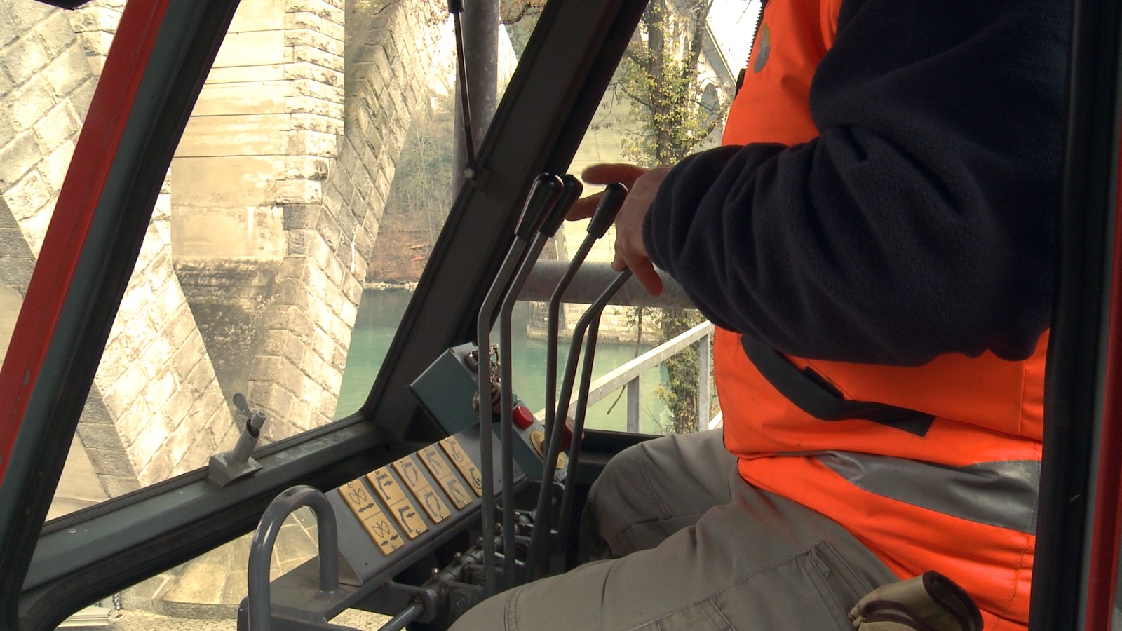 Photogramme : dans une cabine vitrée, un homme en veste orange de chantier tient un levier de commande ; derrière la vitre, une arche de pont en pierre et l'eau verte du fleuve.