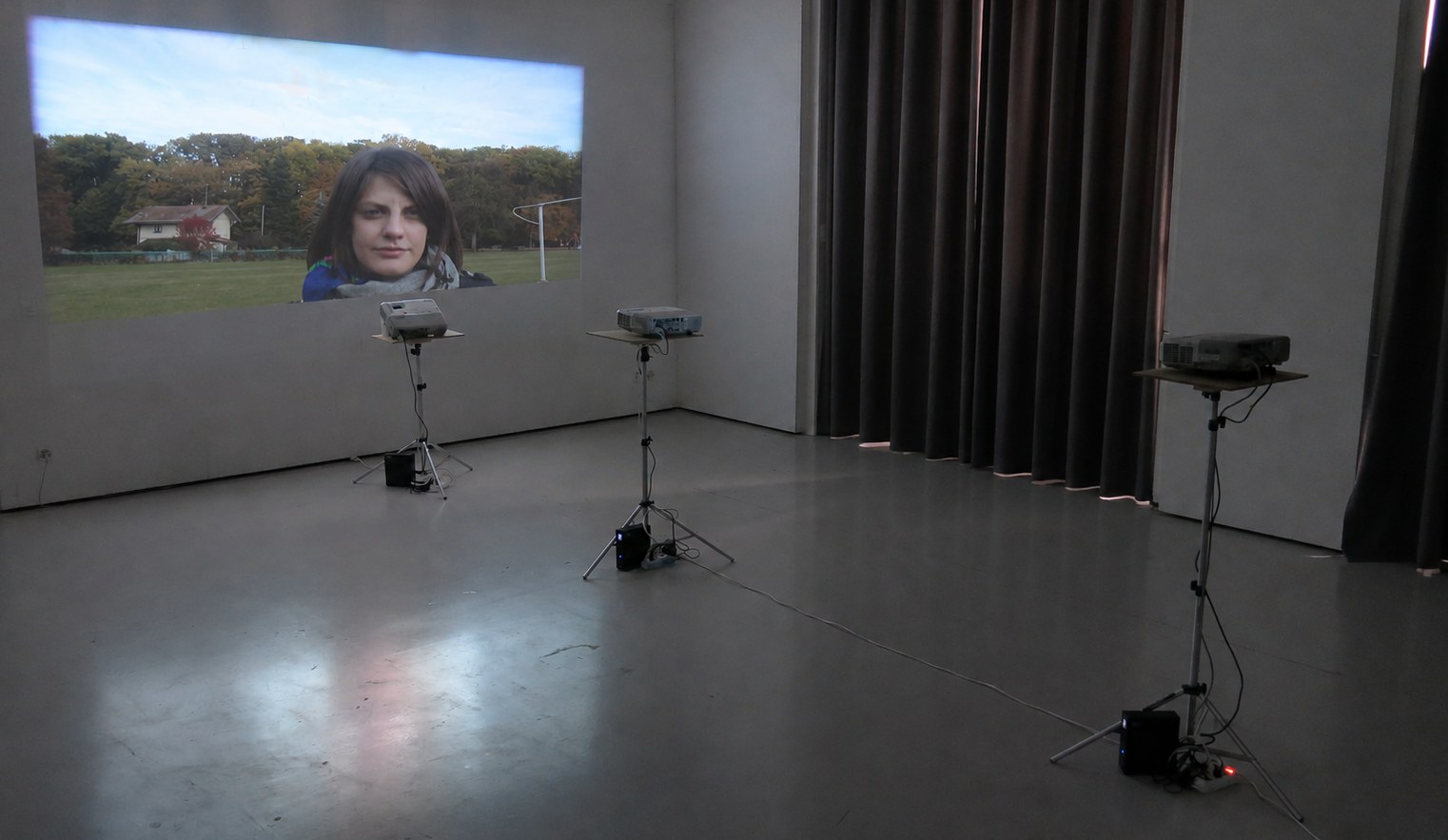 Vue d'installation : dans une salle au sol de béton, un visage projeté en large bandeau sur le mur ; trois projecteurs posés sur des pieds télescopiques, chacun à sa distance.