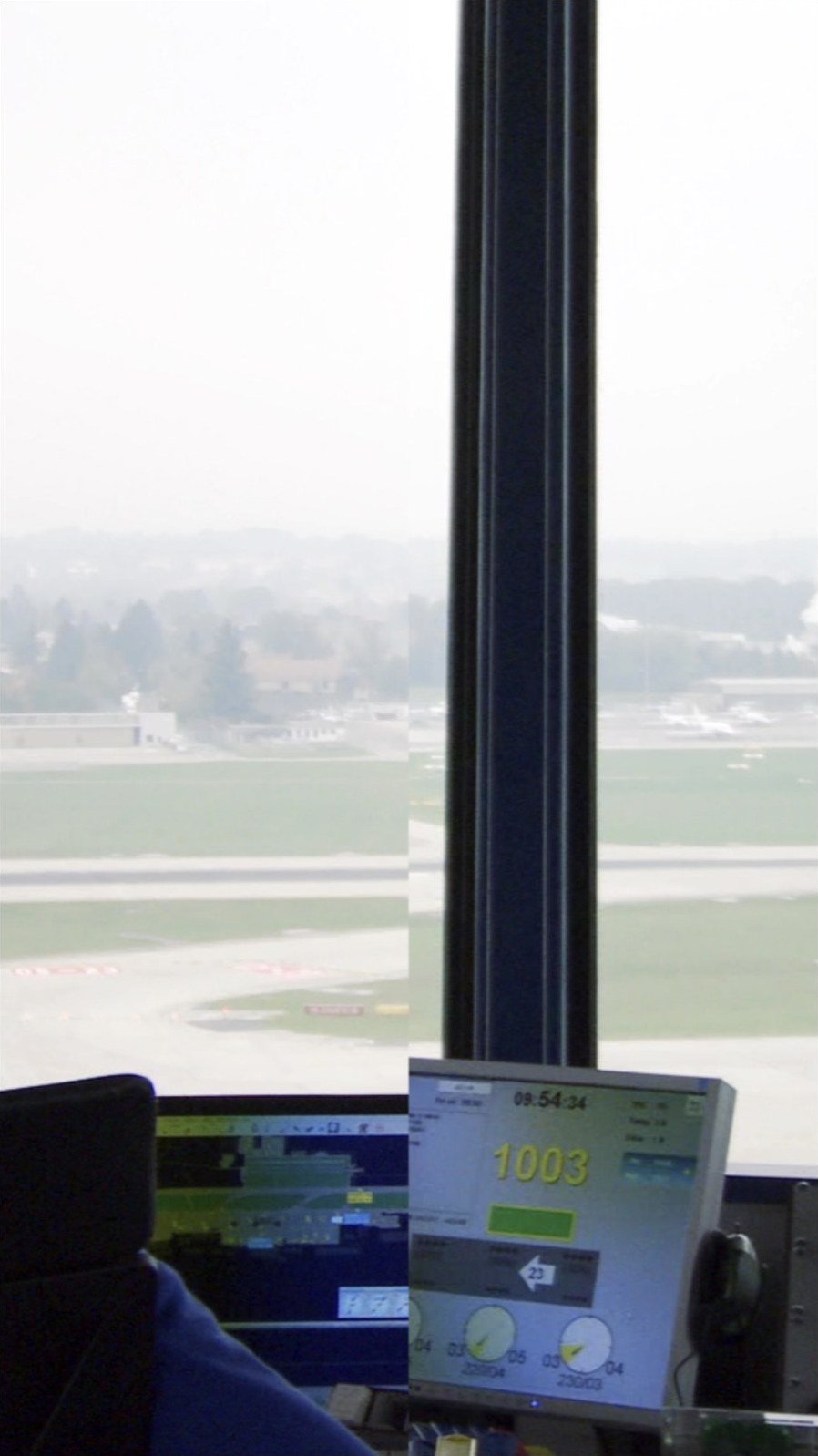Photogramme vertical&nbsp;: vue depuis la tour de contrôle de l'aéroport de Genève. Derrière la vitre, les pistes et le paysage dans une brume claire ; un montant noir de la baie coupe l'image en son milieu. En bas, un écran de contrôle affiche l'heure 09:54:34 et le nombre 1003 en gros caractères jaunes.
