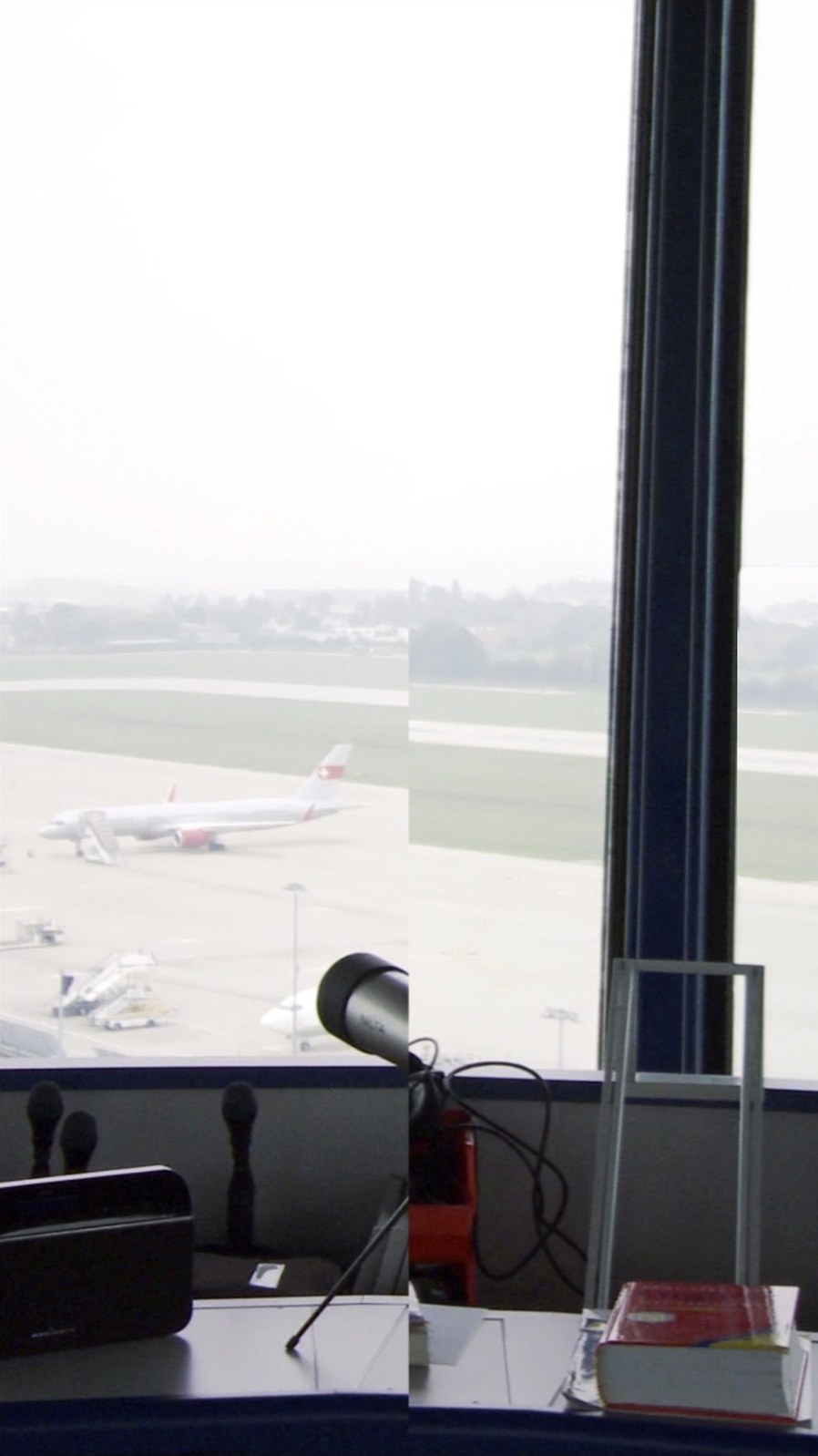 Photogramme vertical&nbsp;: un avion de ligne blanc à la queue rouge stationne sur le tarmac, vu de haut à travers la vitre. La moitié droite de l'image, décalée, ne montre plus que la piste vide et le montant sombre de la baie ; au premier plan, un micro sur pied et un livre posé sur la console.
