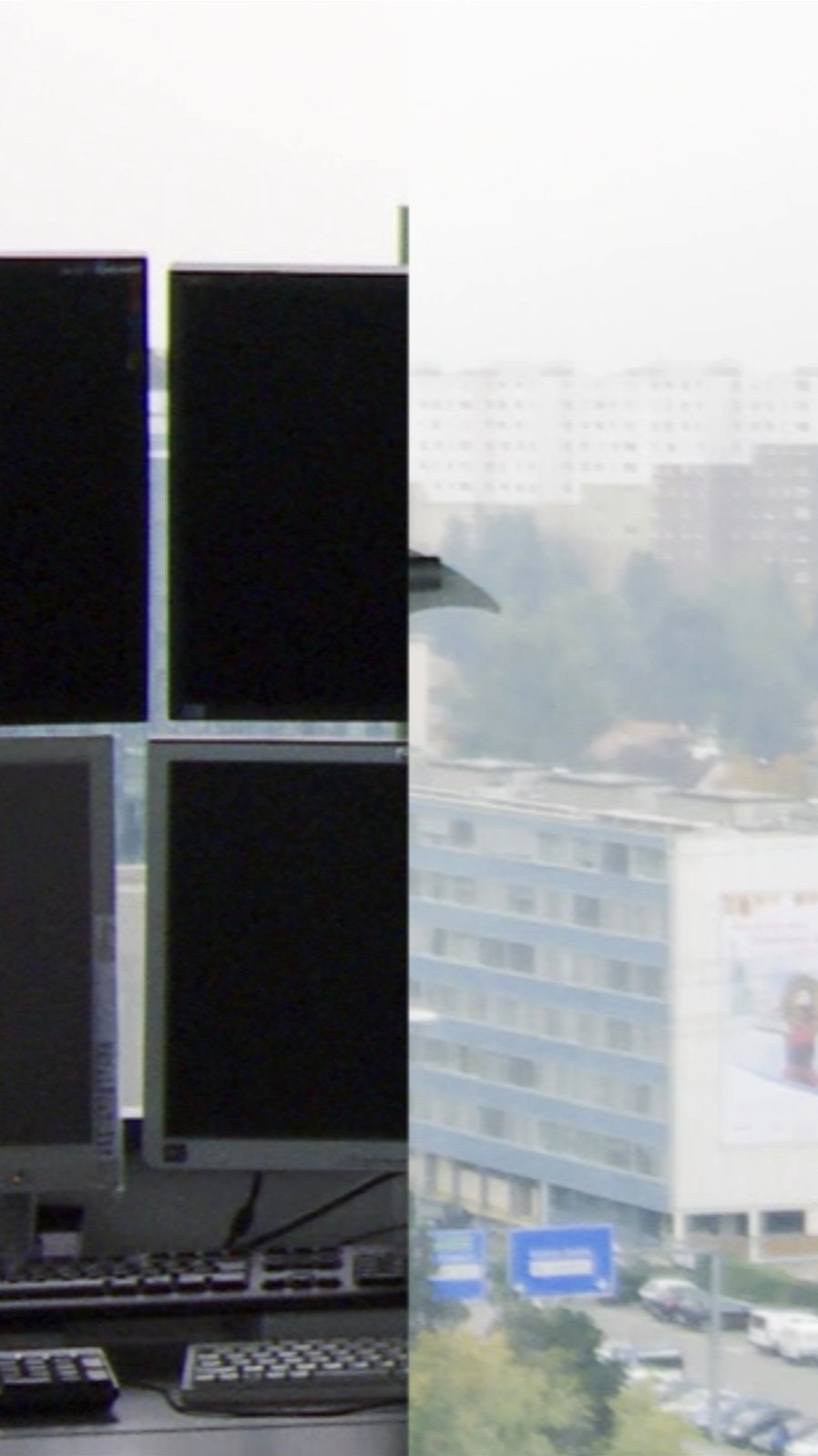 Photogramme vertical&nbsp;: à gauche, quatre moniteurs noirs empilés au-dessus d'un clavier de console ; à droite, la ville vue depuis la tour — immeubles de bureaux, panneaux de signalisation bleus, voitures garées — dans une lumière laiteuse.