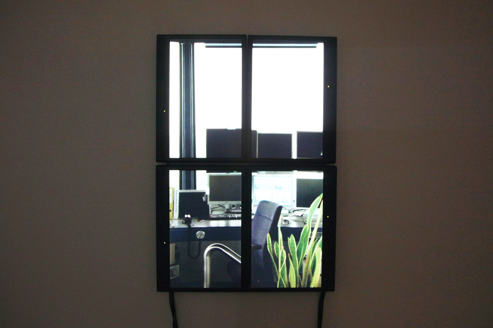 L'œuvre installée&nbsp;: quatre écrans accolés deux par deux forment un rectangle vertical accroché sur un mur sombre, à la manière d'une baie vitrée. Les deux écrans du haut montrent la vitre de la tour à contre-jour, ceux du bas l'intérieur de la salle de contrôle — consoles, fauteuil, une plante grasse au premier plan.