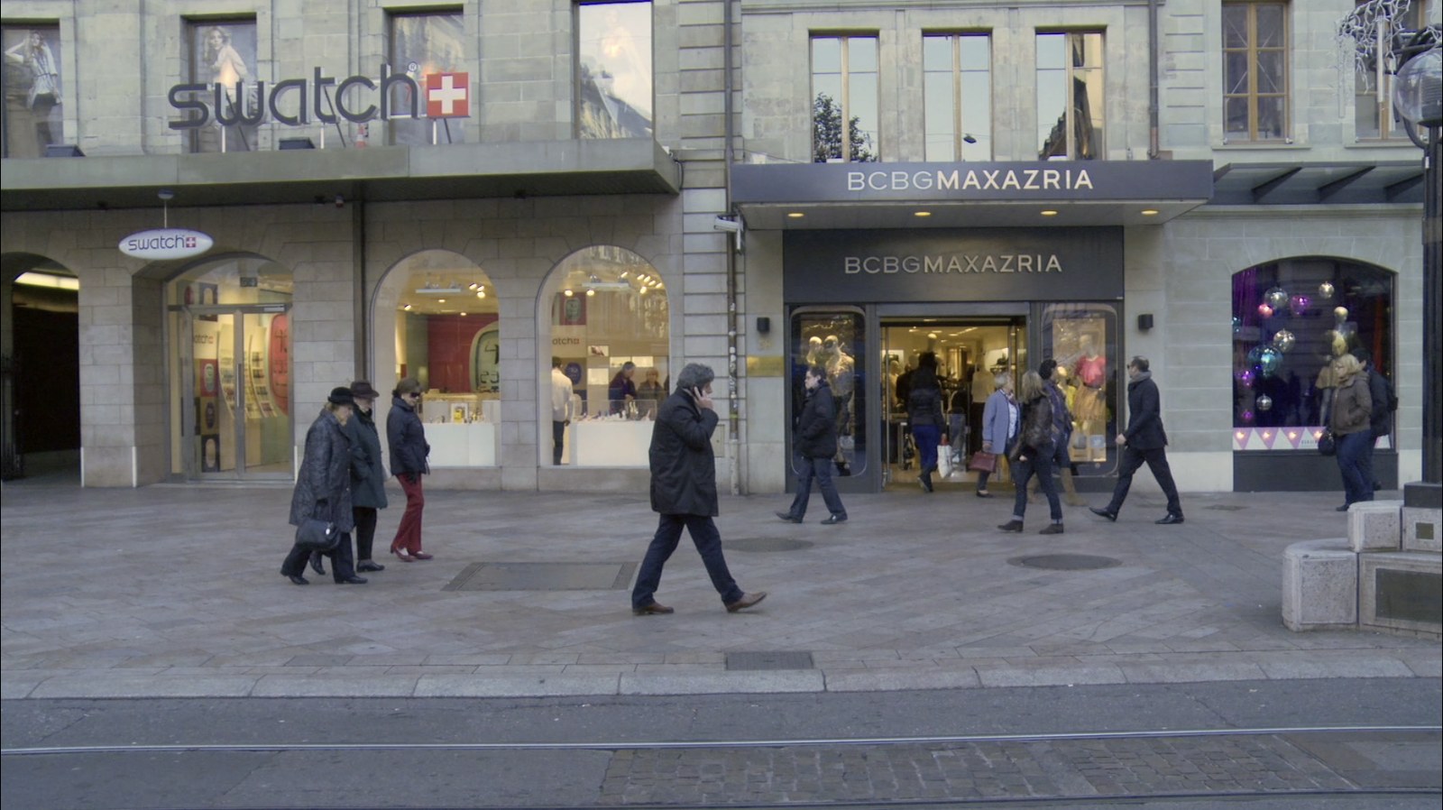Photogramme&nbsp;: une avenue commerçante de Genève à hauteur du regard, encore d'un seul tenant. Devant les vitrines Swatch et BCBG Maxazria, des passants marchent sur le pavé ; au premier plan, les rails du tram.