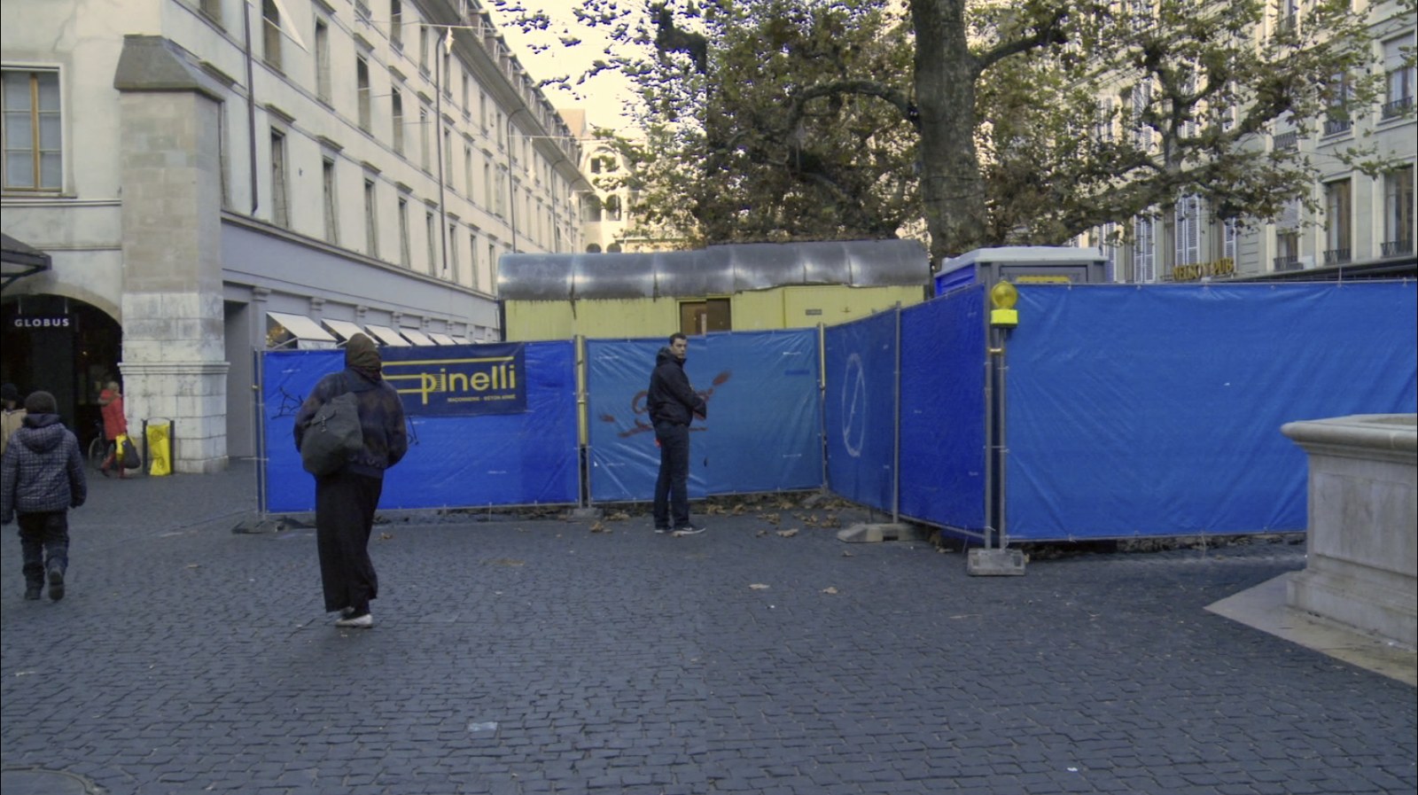 Photogramme&nbsp;: une place pavée bordée d'un chantier ceint de bâches bleues au nom de l'entreprise Pinelli. Un homme au sac à dos traverse le cadre de dos ; à droite, un autre s'est arrêté devant les palissades.