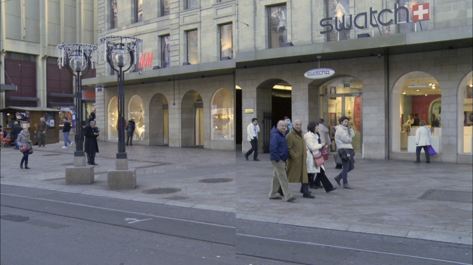 Photogramme&nbsp;: la coupure centrale est désormais nette. À gauche, deux lampadaires devant les arcades d'un grand magasin ; à droite, décalé, un groupe de passants marche devant la boutique Swatch.