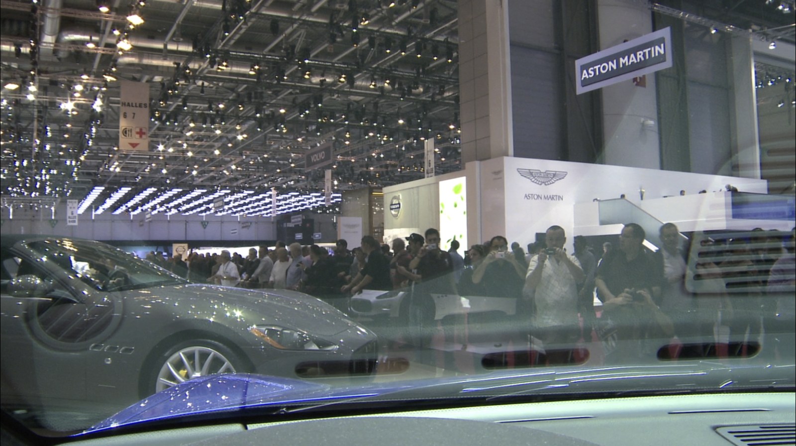 Quatrième photogramme&nbsp;: l'image s'est recomposée et le lieu se révèle. Vue prise depuis l'habitacle d'une voiture, à travers le pare-brise&nbsp;: un coupé gris sur son socle, le stand Aston Martin, la foule massée derrière une barrière et, au plafond, les rampes de projecteurs du hall d'exposition.