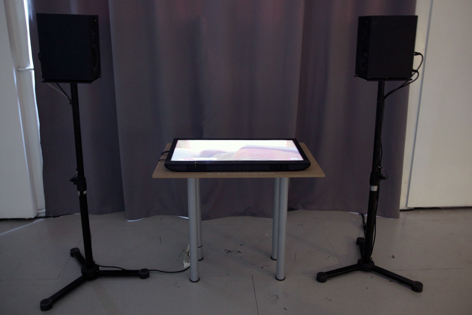 Vue d'installation : un écran plat posé à l'horizontale sur une table basse en bois clair, encadré par deux haut-parleurs noirs montés sur pieds, devant un rideau gris.