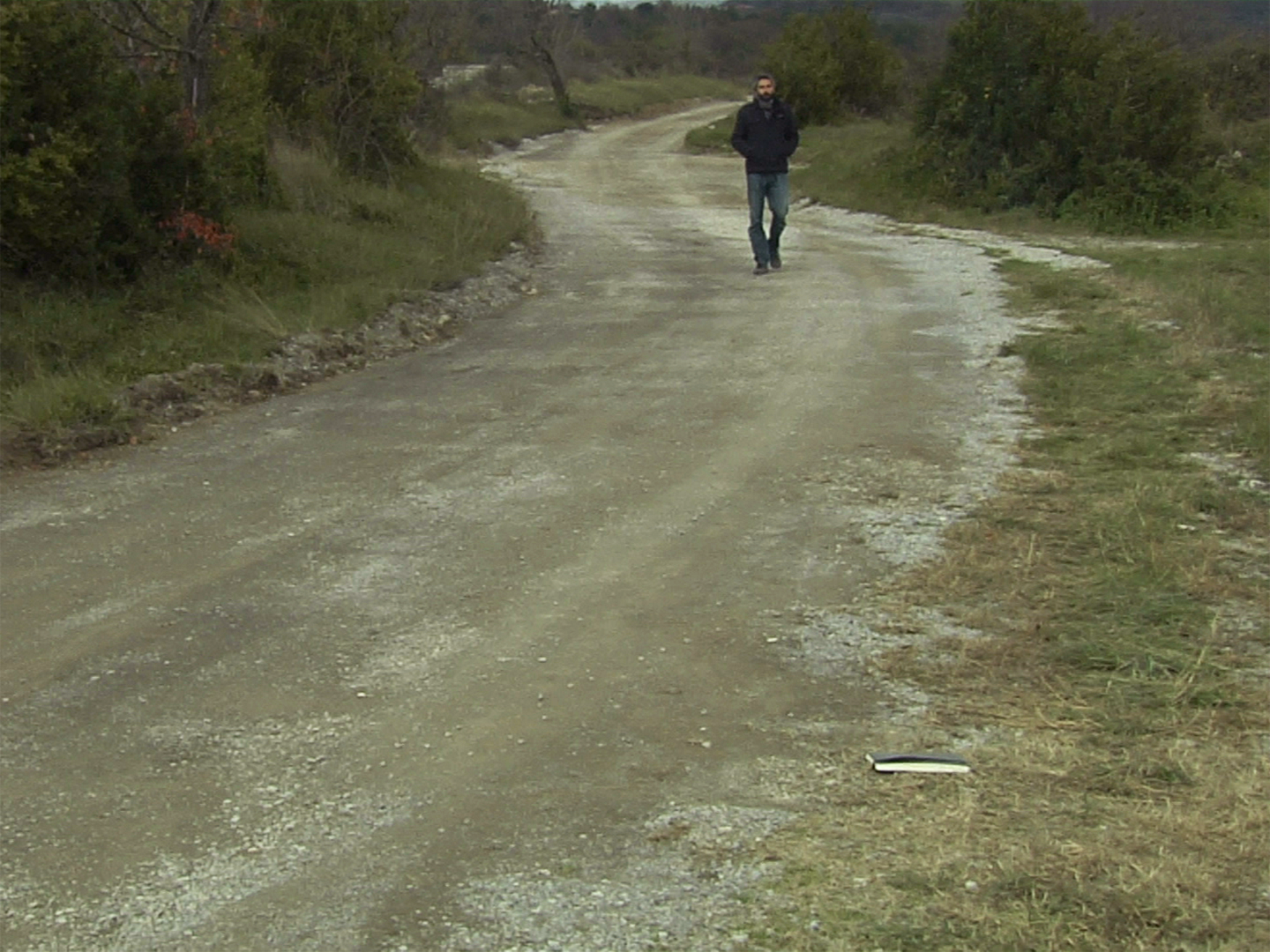 Photogramme : sur un chemin de terre clair bordé de broussailles, un homme en veste sombre marche vers la caméra ; au premier plan, à droite, un carnet ouvert est tombé dans l'herbe.
