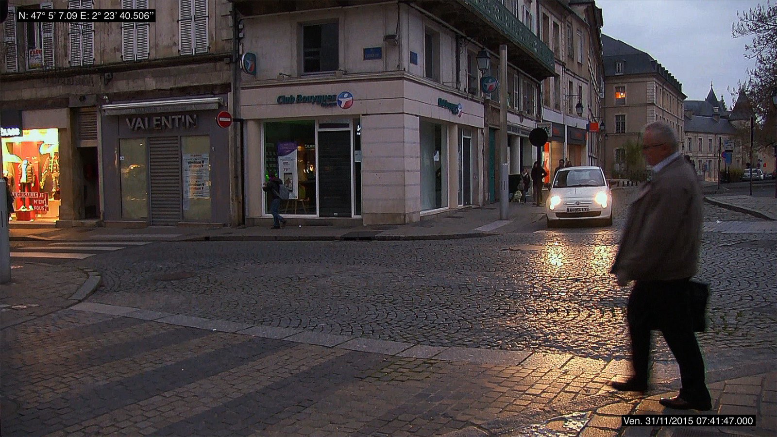Photogramme d'un plan fixe filmé à hauteur de regard, au petit matin&nbsp;: un carrefour pavé de centre-ville, luisant de pluie, bordé d'immeubles clairs et de devantures — une boutique de vêtements éclairée à gauche, une agence de téléphonie à l'angle. Une voiture blanche, phares allumés, attend au débouché de la rue&nbsp;; un homme en veste claire, cartable à la main, traverse au premier plan à droite. En haut à gauche, les coordonnées GPS incrustées&nbsp;; en bas à droite, l'horodatage.