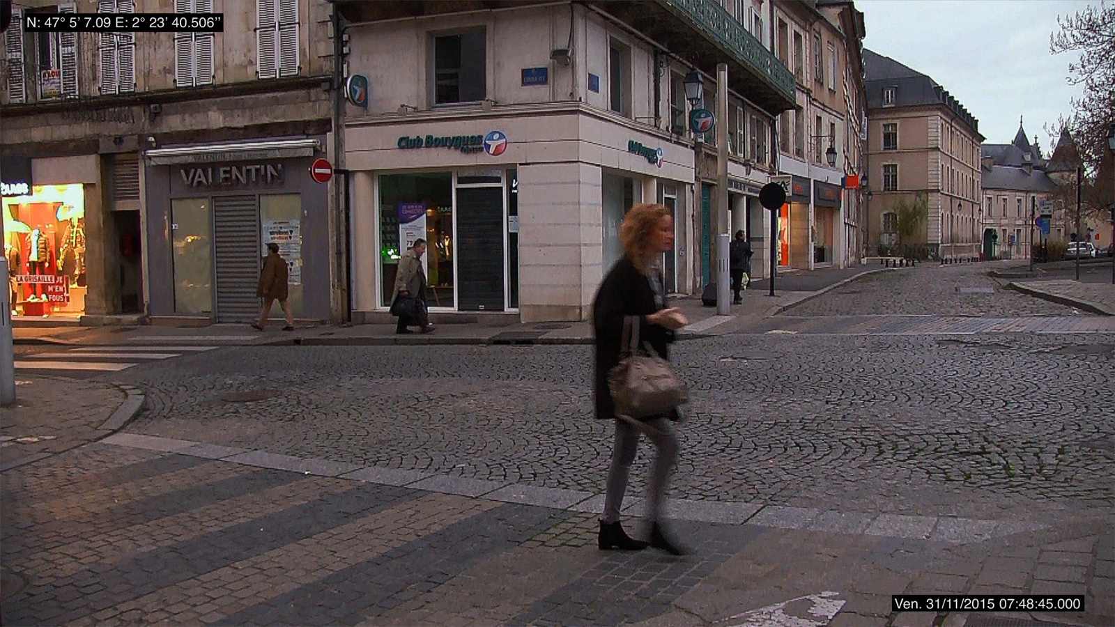 Second photogramme du même plan fixe, un autre jour&nbsp;: le même carrefour pavé, sec cette fois, sous une lumière grise. Une passante en manteau noir, sac au bras, traverse le premier plan de profil&nbsp;; au fond, deux silhouettes s'éloignent le long des devantures et la rue pavée fuit vers la droite, vide. En haut à gauche, les coordonnées GPS incrustées&nbsp;; en bas à droite, l'horodatage.
