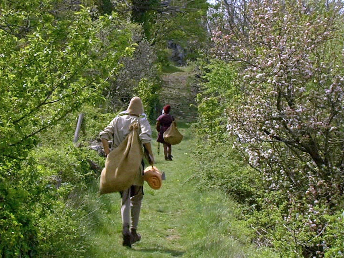 Photogramme : sur un sentier herbeux bordé d'aubépines en fleurs, deux personnes en costume du XVIIᵉ siècle s'éloignent, un ballot de toile sur l'épaule.