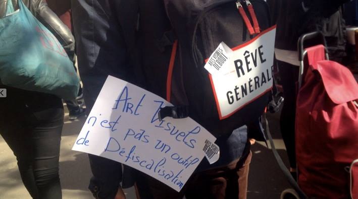 Photogramme : dans une manifestation, une pancarte manuscrite « Art visuels n'est pas un outil de Défiscalisation » portée devant un sac à dos, à côté d'un dossard « Rêve général ».