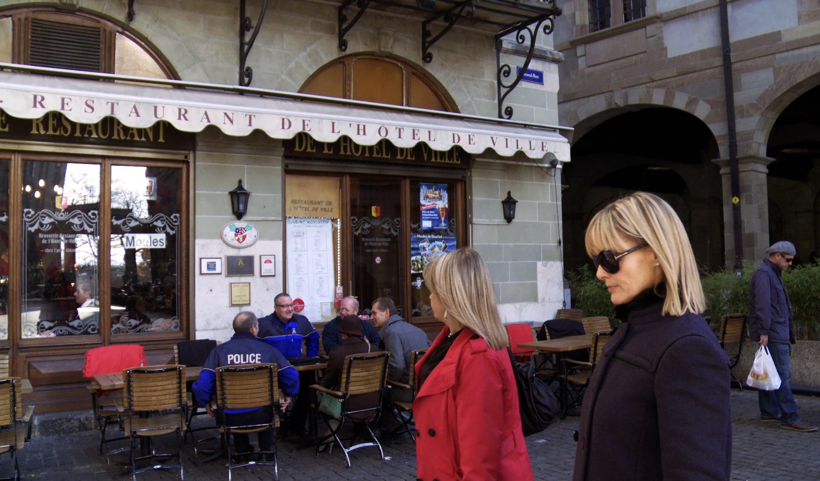 Volet gauche du diptyque, la photographie d'origine&nbsp;: la terrasse d'une brasserie sur une place pavée, store à festons portant «&nbsp;RESTAURANT DE L'HÔTEL DE VILLE&nbsp;». Autour des tables en bois, des consommateurs attablés, dont un homme en blouson bleu marqué POLICE, de dos. Au premier plan, deux femmes traversent le cadre, l'une en manteau rouge, l'autre en manteau sombre et lunettes noires&nbsp;; à droite, un passant s'éloigne sous les arcades, un sac à la main.