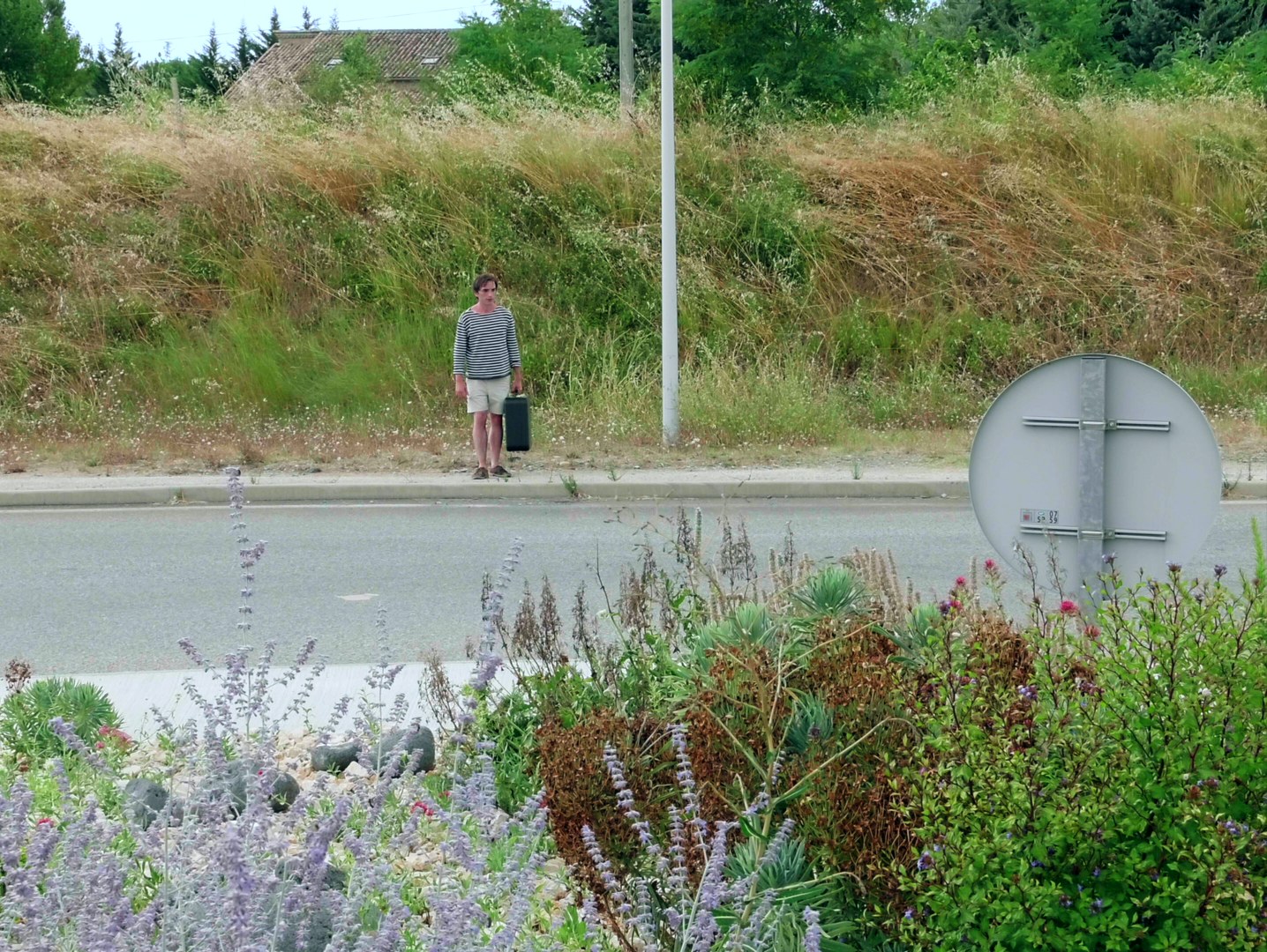 Photogramme : au bord d'une route, devant un talus d'herbes sèches, un homme en marinière et short attend, une mallette noire à la main ; à droite, le dos d'un panneau de signalisation.