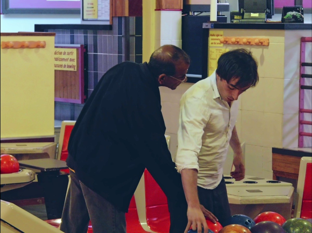 Photogramme : dans un bowling aux couleurs vives, un homme âgé penché ramasse une boule tandis qu'un homme jeune en chemise claire, debout, regarde vers le sol.