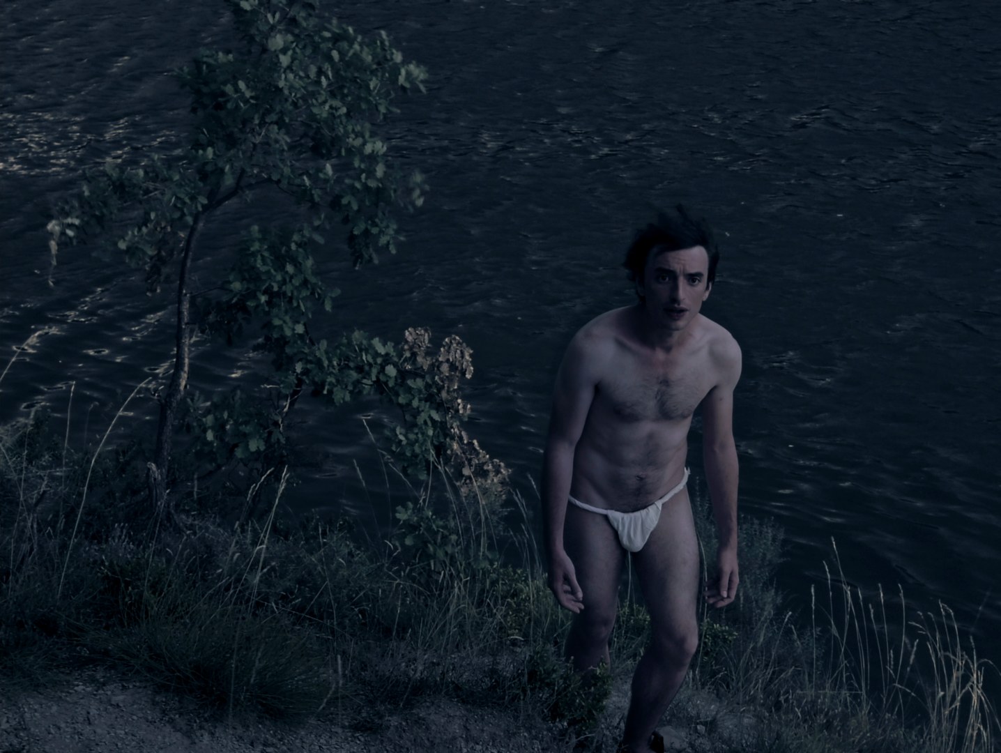 Photogramme nocturne : au bord d'une eau sombre, un homme torse nu, en linge blanc, se tient debout dans les herbes et fixe l'objectif.
