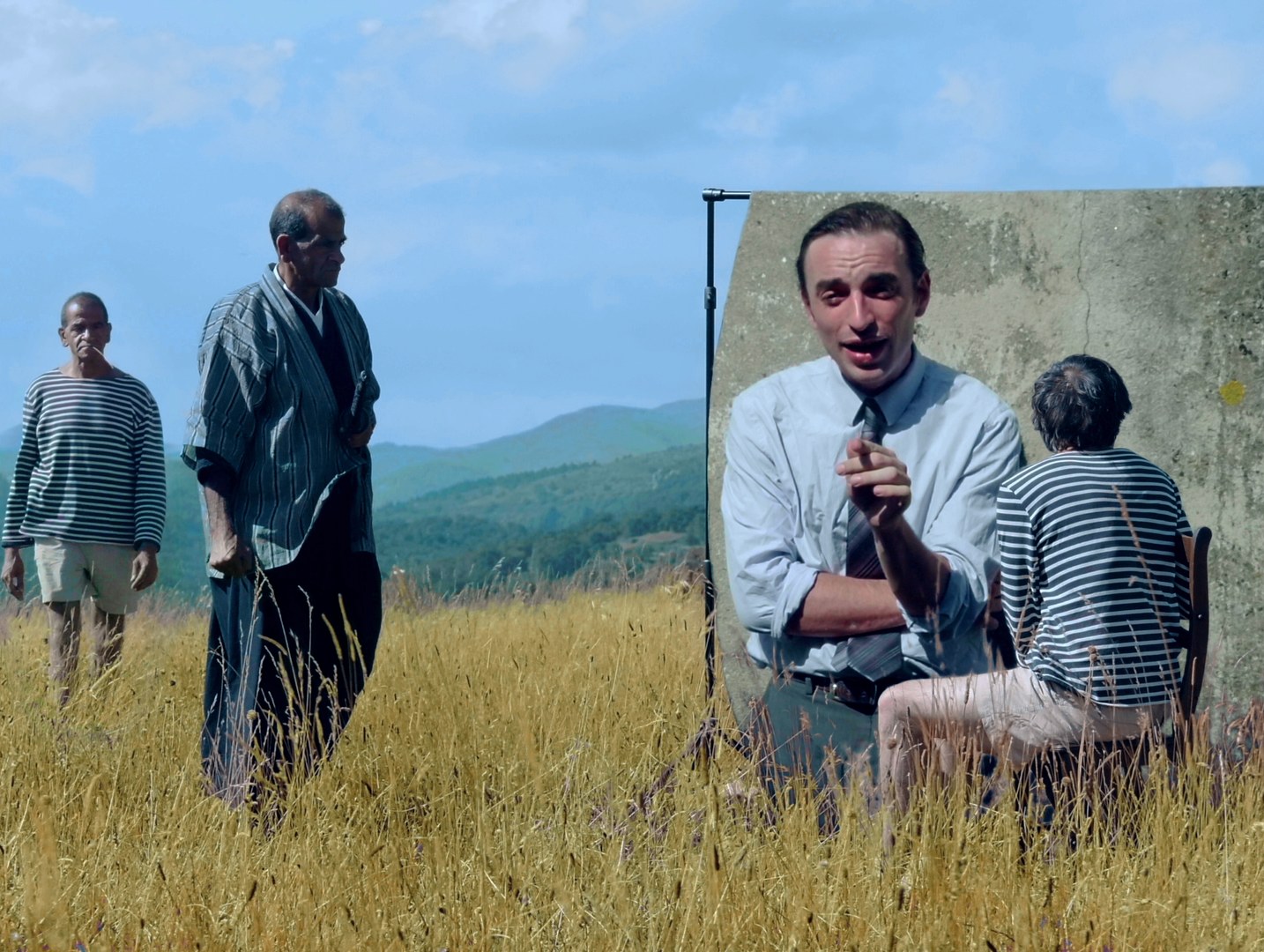 Photogramme : dans un pré d'herbes hautes, deux hommes debout — l'un en marinière, l'autre en kimono rayé ; à droite, contre un mur de béton, un homme en chemise et cravate parle, assis face à une personne de dos.