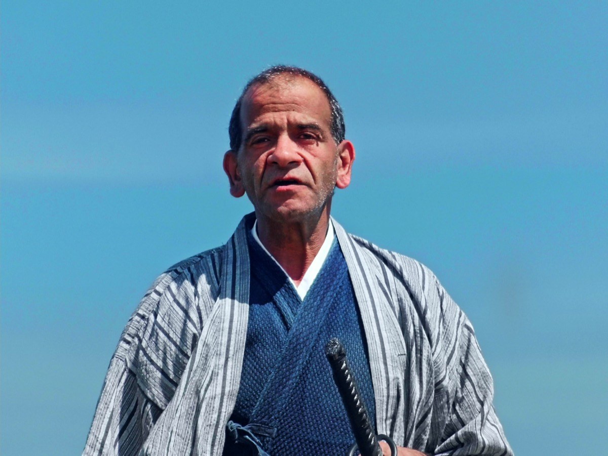 Photogramme : sur fond de ciel, le buste d'un homme en kimono rayé et gilet bleu, un sabre glissé à la ceinture ; il parle.