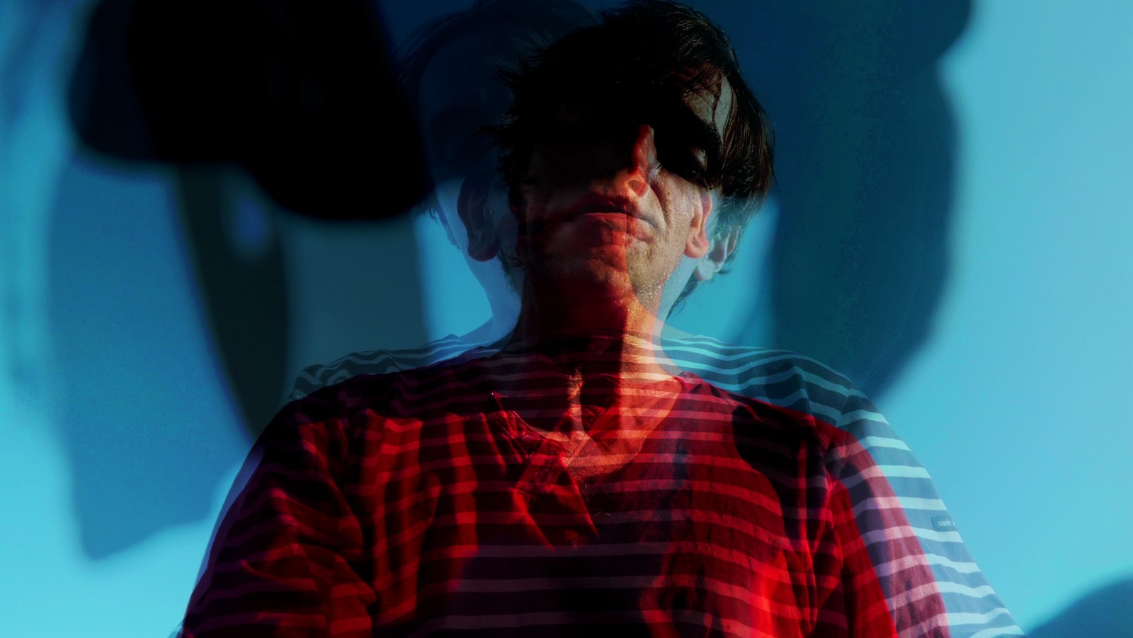 Photogramme en surimpression : deux visages se superposent sur un fond bleu, l'un teinté de rouge, l'autre en marinière rayée ; leurs ombres se projettent au mur.