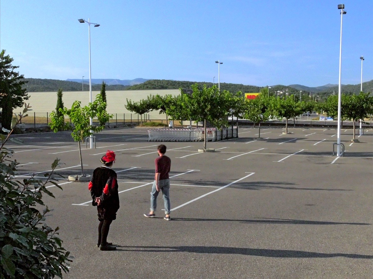 Photogramme : sur le parking désert d'une zone commerciale, deux hommes se tiennent à distance l'un de l'autre — l'un en costume rouge et noir à coiffe emplumée, l'autre en pull rayé.
