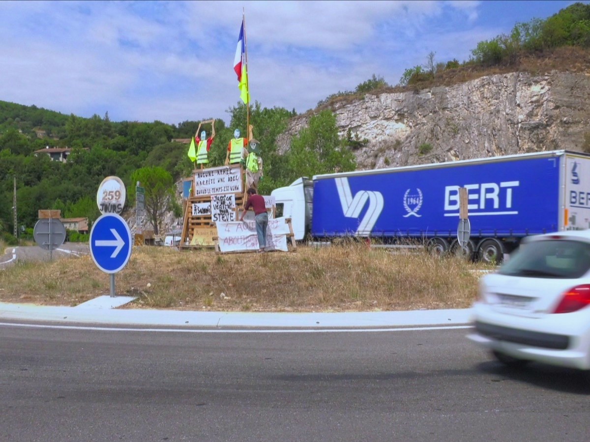 Photogramme : sur un rond-point, une cabane de gilets jaunes couverte de pancartes et surmontée d'un drapeau ; un camion et une voiture passent, un panneau annonce « 259 jours ».