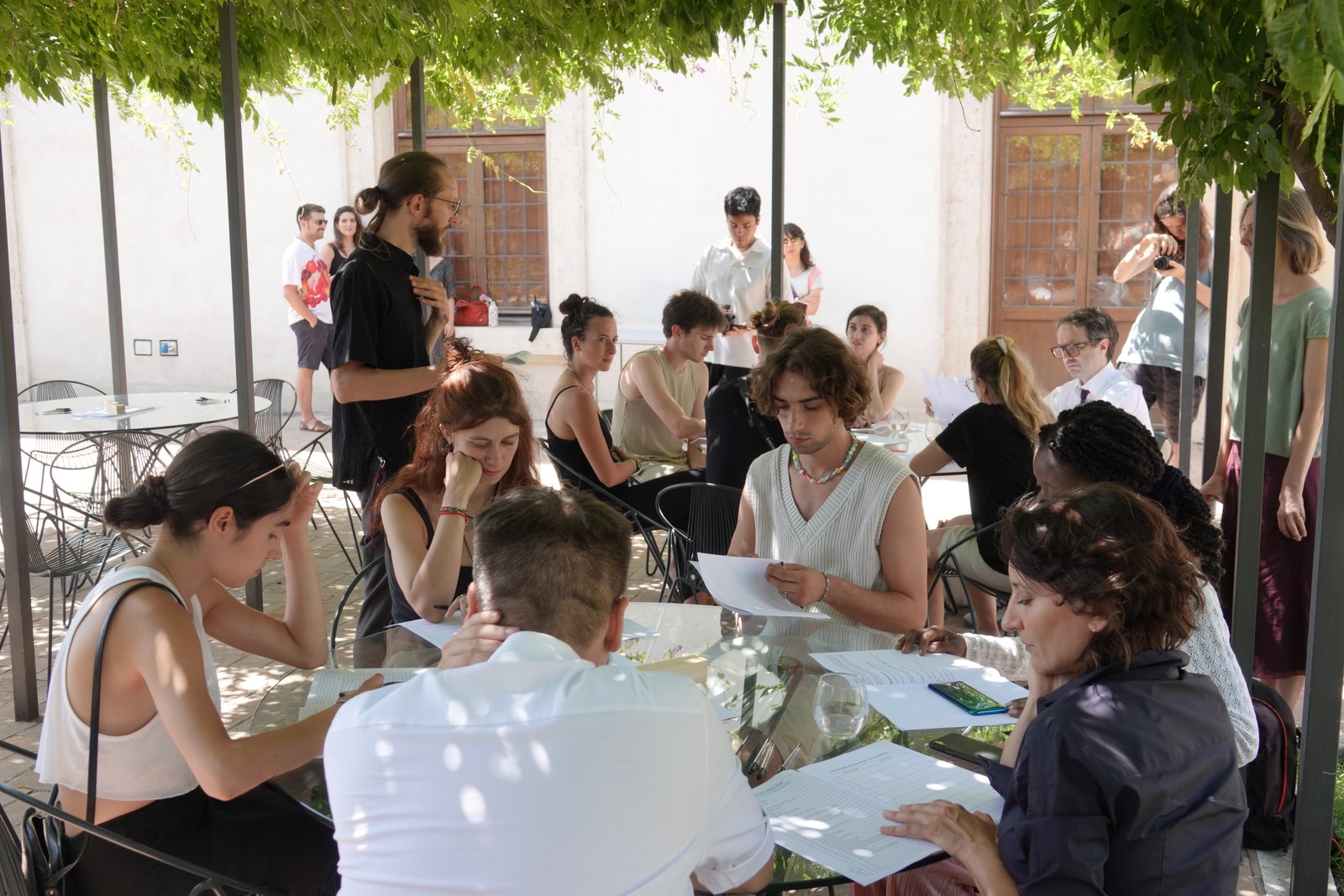 La performance en cours : sous une treille de glycine, des participants penchés sur des feuilles autour de tables rondes en verre, d'autres debout en arrière-plan, dans une cour aux murs clairs.