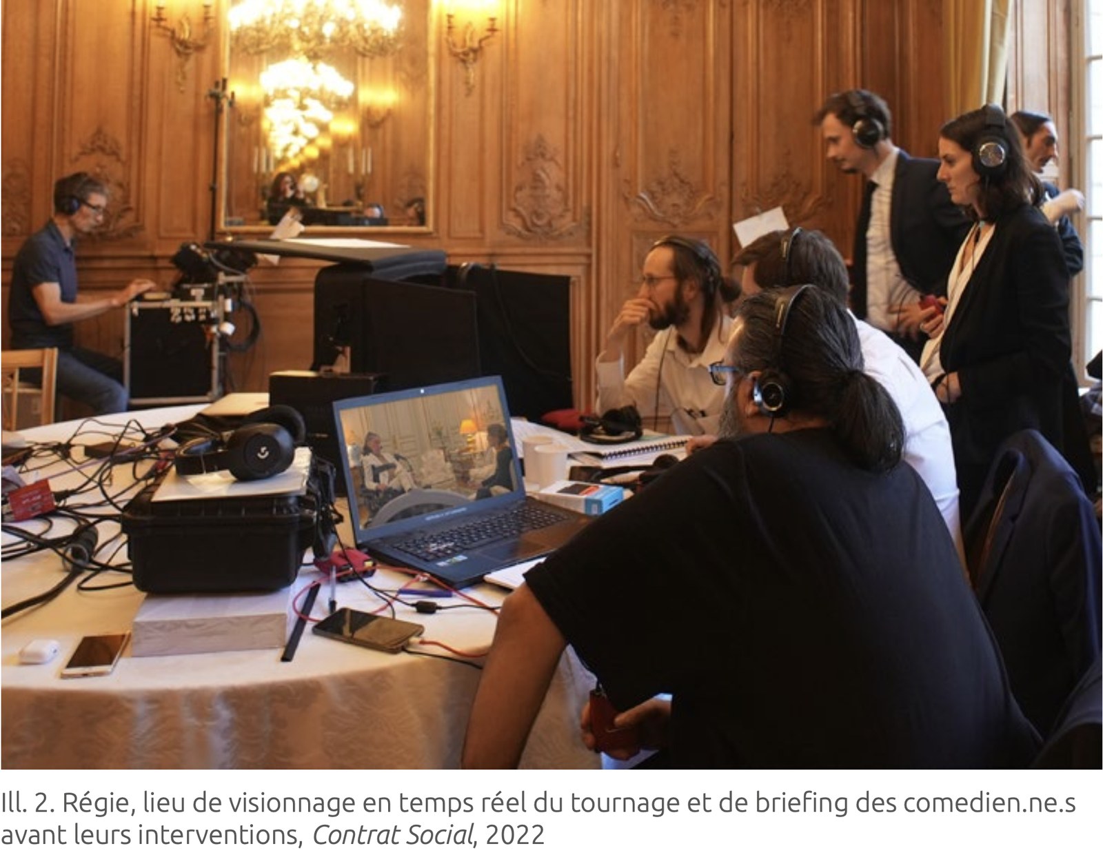 Vue de la régie, dans une salle aux boiseries dorées&nbsp;: autour d'une table nappée encombrée de câbles, de casques et de caisses, une dizaine de personnes casquées suivent le tournage sur un ordinateur portable où se distingue la scène du salon. En arrière-plan, un opérateur près d'une caméra sur pied, un grand miroir et un lustre allumé. Une légende incrustée sous l'image désigne la régie comme lieu de visionnage en temps réel et de briefing des comédiennes et comédiens, Contrat Social, 2022.