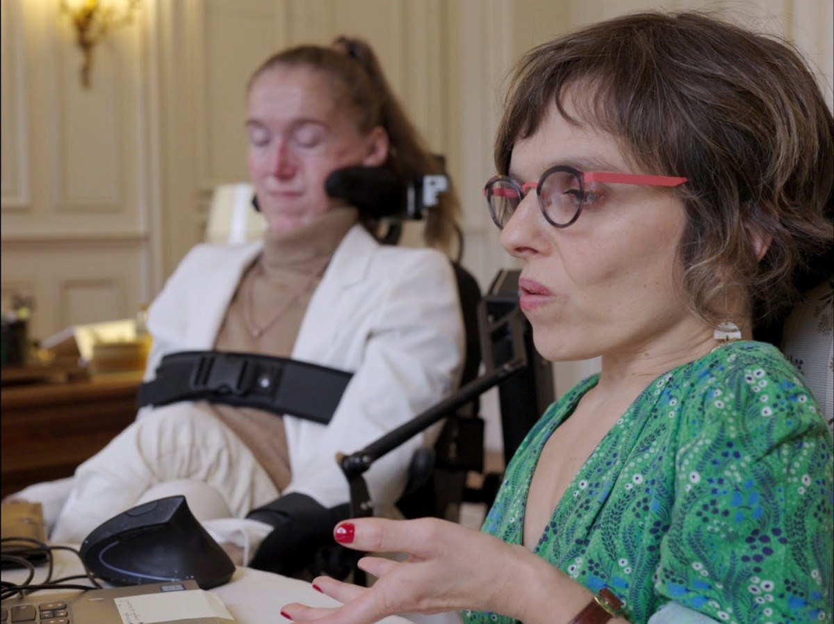 Photogramme : dans un salon lambrissé, une femme en fauteuil roulant, veste blanche et appui-tête, écoute ; au premier plan, une autre femme aux lunettes cerclées de rouge parle en gesticulant.