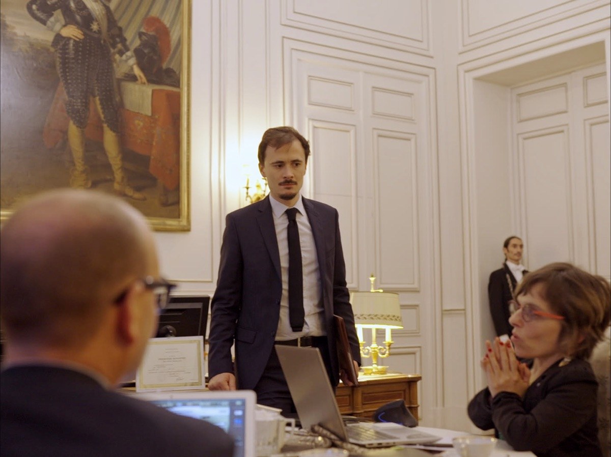 Photogramme : un homme en costume sombre se tient debout derrière un bureau, dans un salon lambrissé de blanc où pend le grand portrait peint d'un dignitaire ; deux personnes l'écoutent au premier plan.