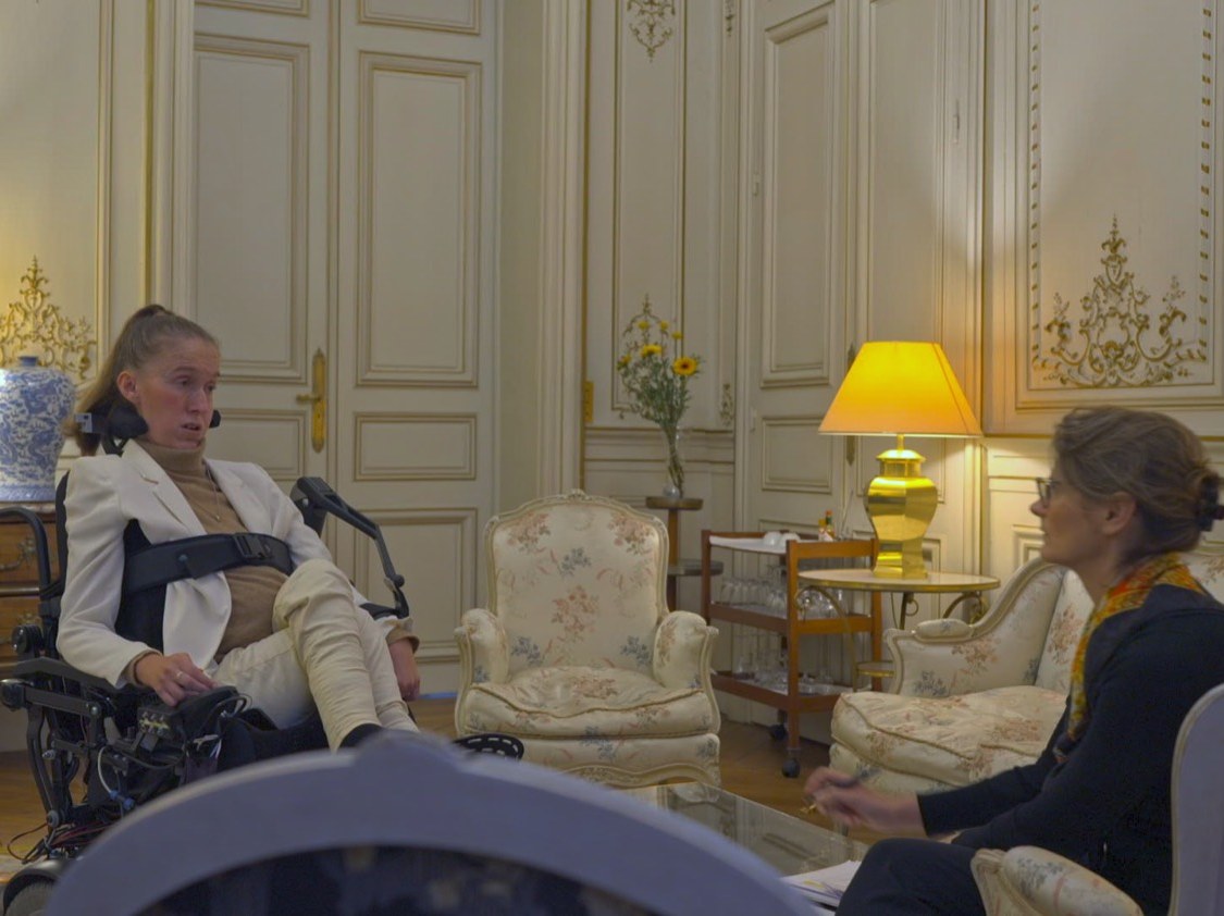 Photogramme : plan large d'un salon doré meublé de fauteuils fleuris ; à gauche, une femme en fauteuil roulant s'adresse à une autre femme assise à droite.