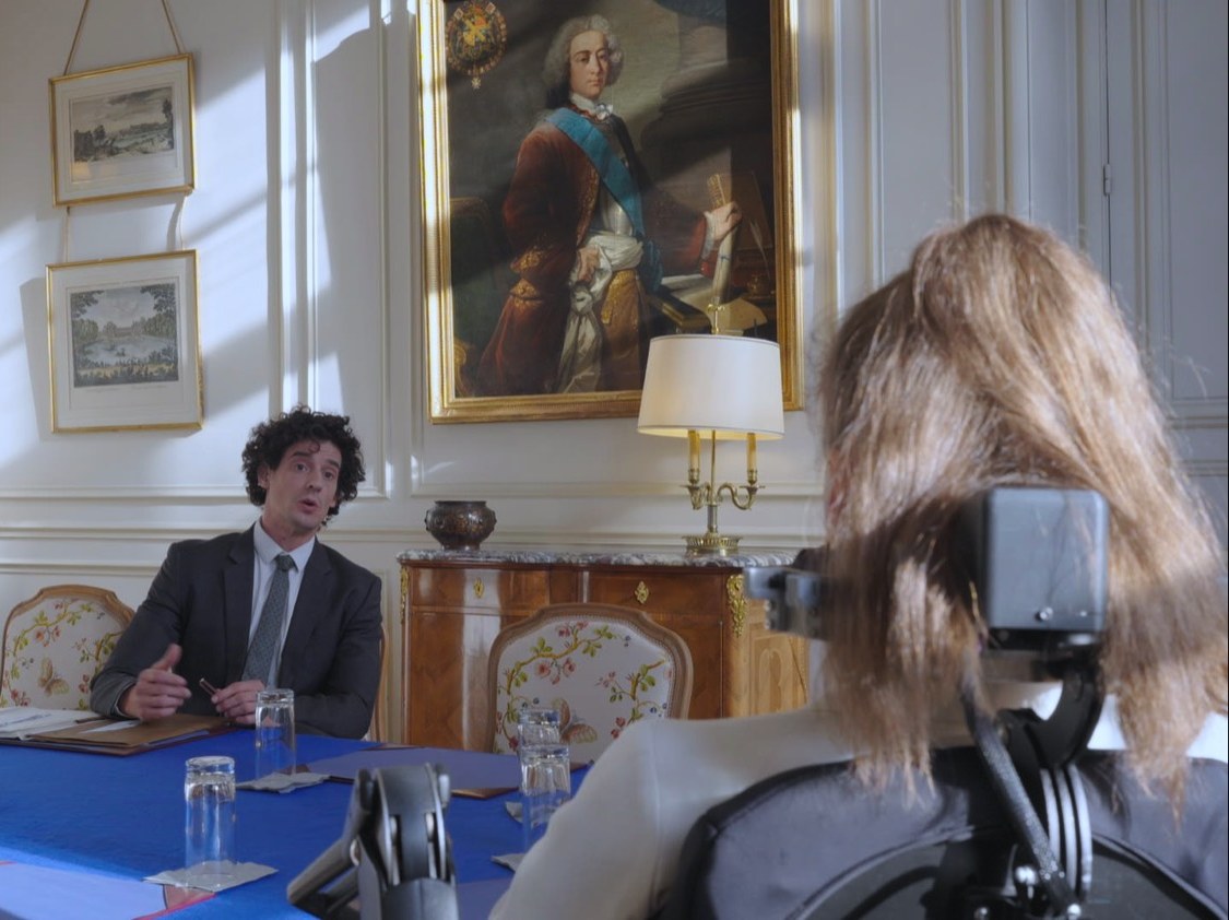 Photogramme : autour d'une table de réunion couverte d'un tapis bleu, un homme aux cheveux bouclés parle ; au premier plan, de dos, une personne en fauteuil roulant l'écoute, sous un portrait peint du XVIIIᵉ siècle.