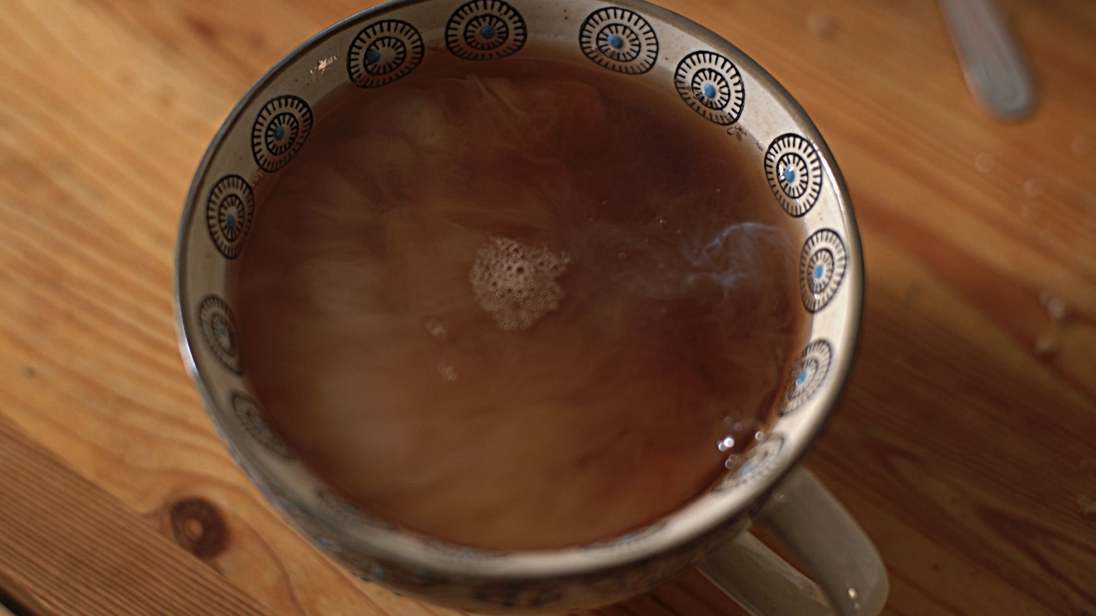 Photogramme : vu de haut, un bol de thé au lait posé sur une table de bois clair ; la vapeur s'enroule à la surface, le bord du bol est orné de rosaces bleues.