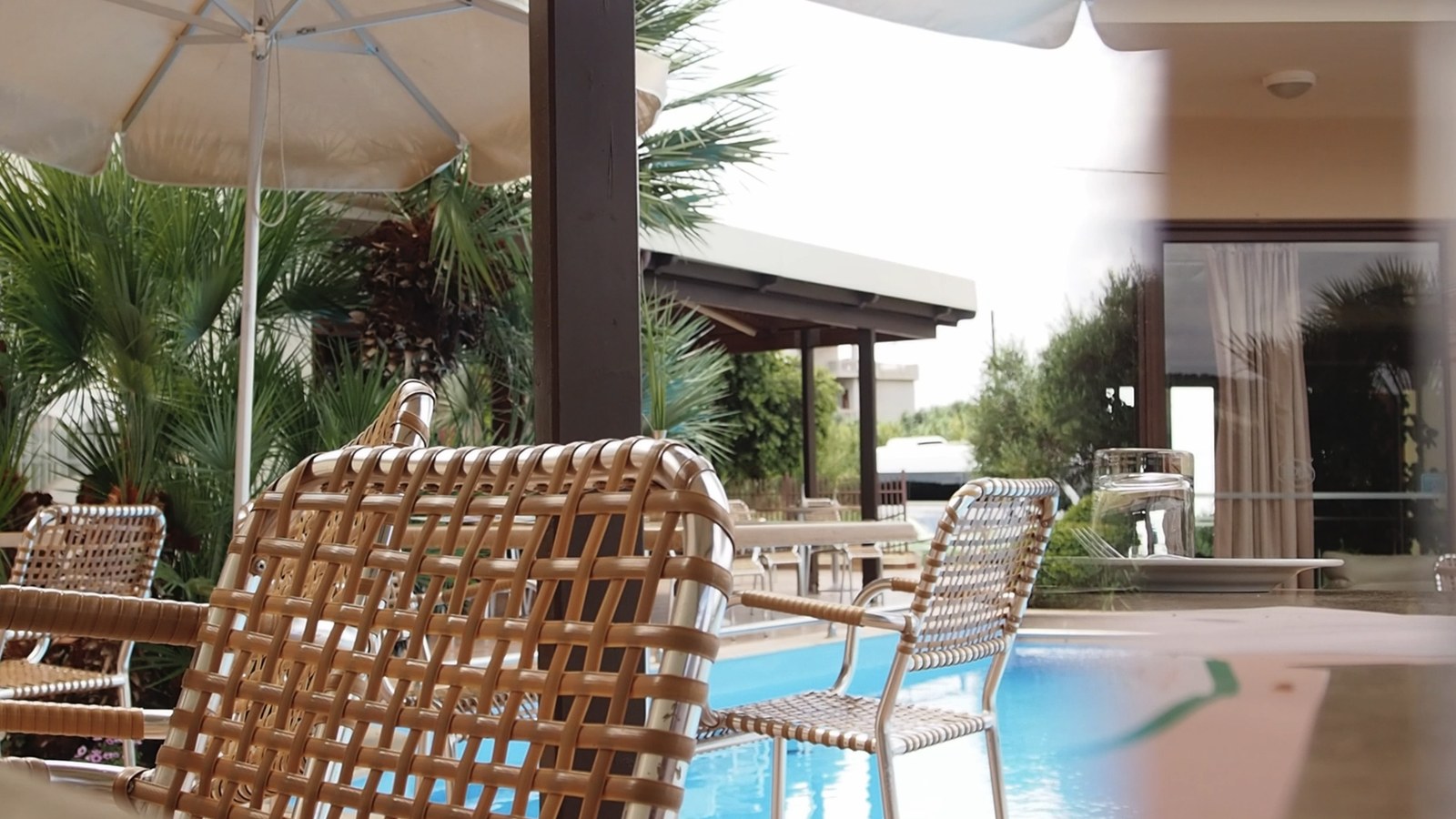 Photogramme : au bord d'une piscine d'hôtel, des fauteuils de résine tressée sous un parasol blanc, des palmiers et une baie vitrée derrière.