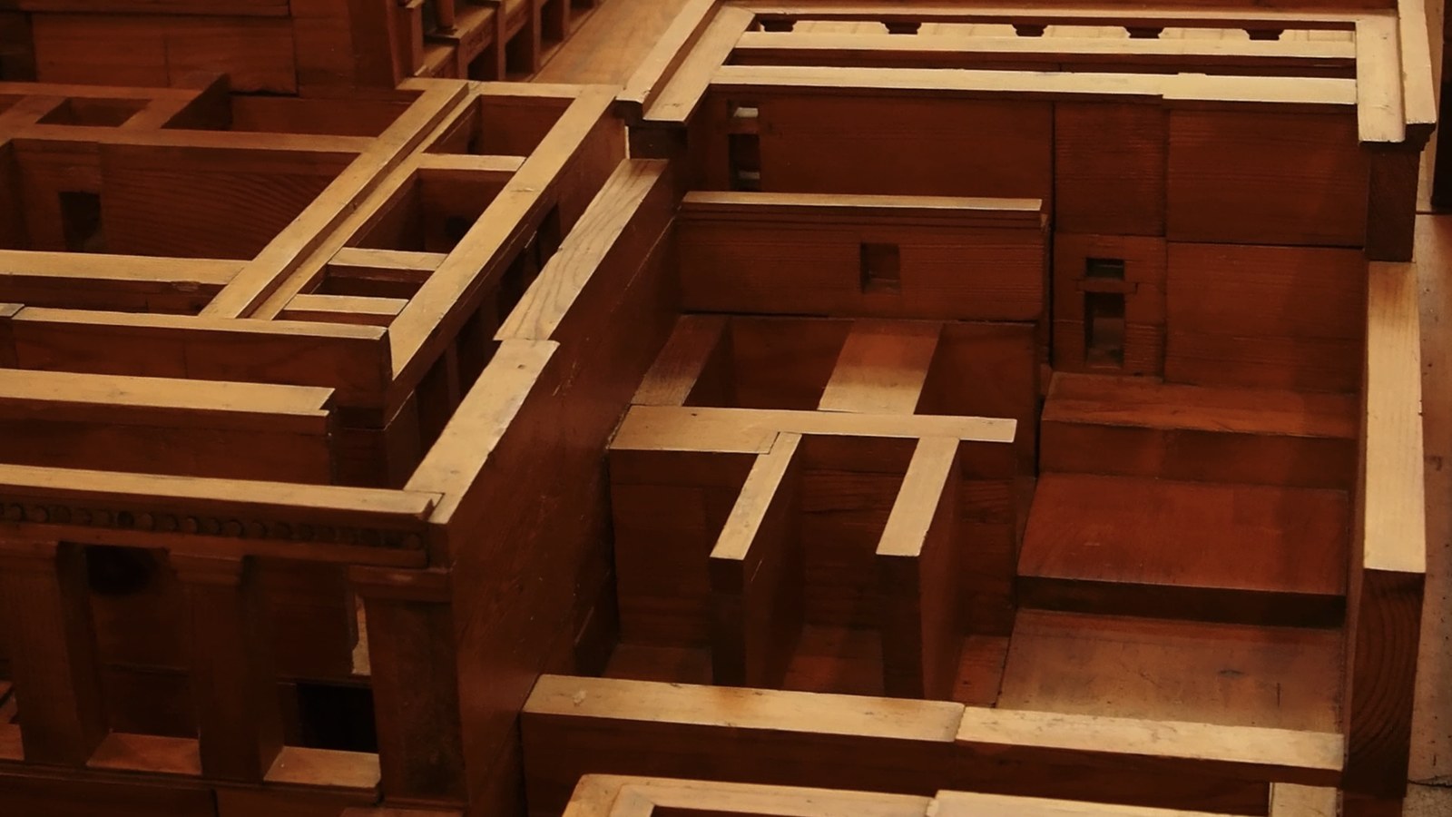 Photogramme : la maquette en bois d'un palais antique, vue de haut — murs, escaliers et cloisons emboîtés, en deux essences claire et sombre.